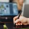pexels photo 5904063 5904063 Close-up of a person using a stylus pen on a digital pad with a blurred laptop background.